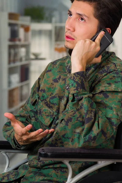 Portrait of a handsome young soldier sitting on wheel chair using his cellphone, in a blurred background — Stock Photo, Image