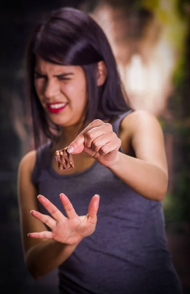 Mooie en eenzaam meisje lijden complet, holding in haar hand een stukje chocolade en doen een teken van stop met haar andere hand, in een onscherpe achtergrond — Stockfoto