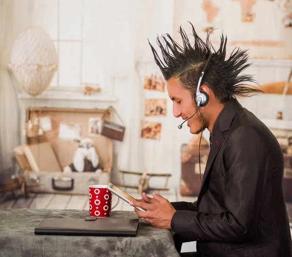 Primer plano del hombre punk de la oficina, con un traje con un peinado de cresta, usando su tableta y auriculares para trabajar en la oficina, en un fondo borroso —  Fotos de Stock