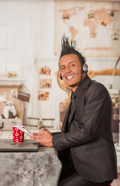Крупним планом усміхнений офісний панк-чоловік, одягнений в костюм зі стилем гребеня волосся, використовуючи свій планшет і навушники для роботи в офісі, на розмитому фоні — стокове фото