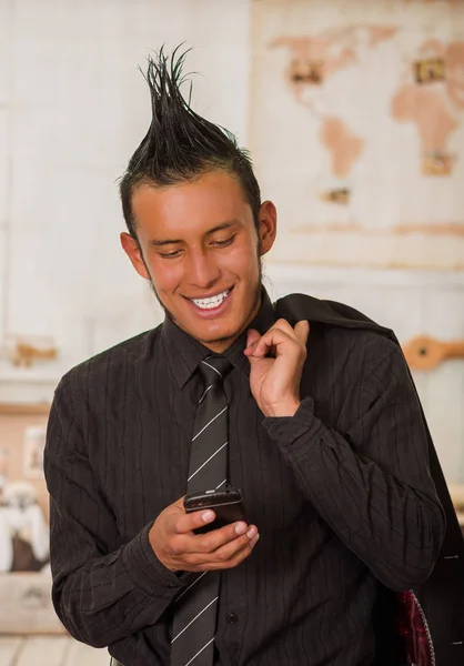 Close up of happy office punk worker wearing a suit with a crest, holding with one hand his jacket in the shoulder and using his cellphone in the office in a blurred background — Stock Photo, Image
