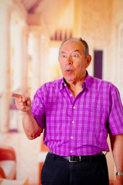 Bir yerde bir bulanık arka planda mor bir kare t-shirt giyen için eliyle işaret mutlu bir yaşlı adamın yakın çekim