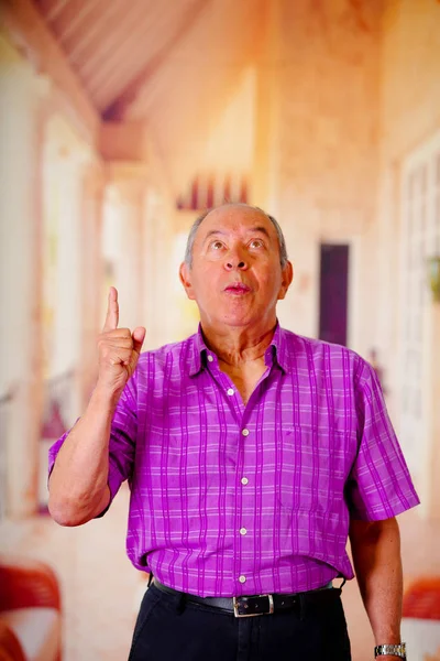 Крупным планом счастливого старика, показывающего рукой вверх и одетого в фиолетовую квадратную футболку на размытом фоне — стоковое фото