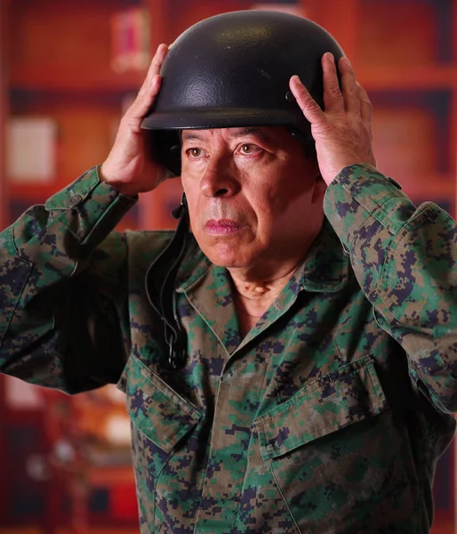 Gros plan d'un vétéran sérieux portant un casque et un uniforme militaire, les deux mains ajustant le casque, dans un arrière-plan flou — Photo