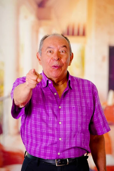 Primer plano de un anciano feliz, señalando con la mano al frente y llevando una camiseta cuadrada púrpura en un fondo borroso — Foto de Stock