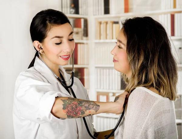 Beautiful tattooed young doctor using a stestoscope checking the chest of the patient, in office background — Stock Photo, Image