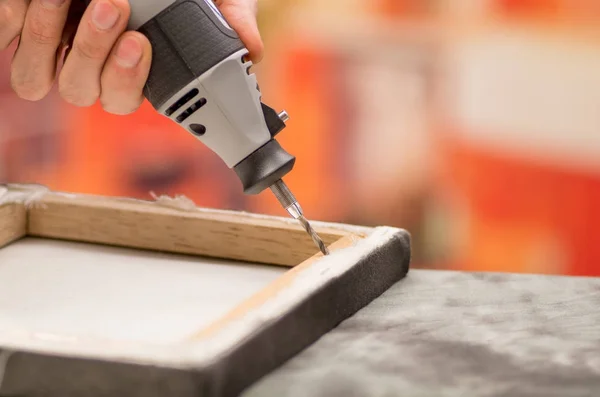 Close-up de um homem trabalhador perfurando uma moldura de madeira com sua broca sobre uma mesa cinza em um fundo embaçado — Fotografia de Stock