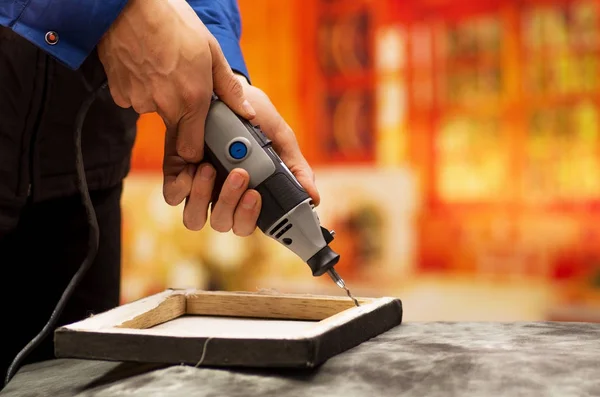 Nahaufnahme eines fleißigen Mannes, der mit seinem Bohrer einen Holzrahmen bohrt, mit Bohrzubehör über einem grauen Tisch vor verschwommenem Hintergrund — Stockfoto