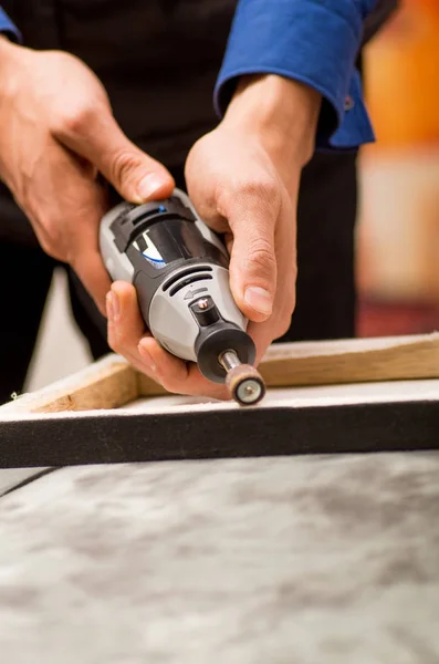 Close-up de um homem trabalhador usando um polidor em uma moldura de madeira, em uma mesa cinza em um fundo desfocado — Fotografia de Stock