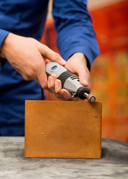 Closeup of a hardworker man wearing a blue t-shirt and using a polisher in a piece of wood, on a gray table in a blurred background — Stock Photo, Image