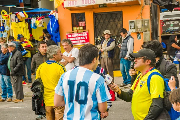 QUITO, ECUADOR - 11 DE OCTUBRE DE 2017: Primer plano del reportero conversando con algunos aficionados argentinos que rodean a una multitud de personas con camisa de fútbol oficial, gritando y saltando apoyando a su equipo — Foto de Stock