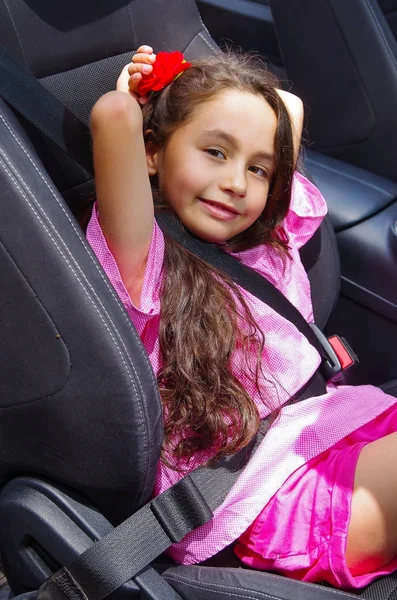 Close up of little beatiful girl sitting in the car weaing a pink dress and a red flower in her head using a safety belt — Stock Photo, Image