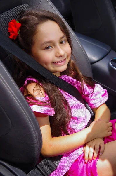 Close up of little beatiful girl sitting in the car wearing a pink dress and a red flower in her head using a safety belt