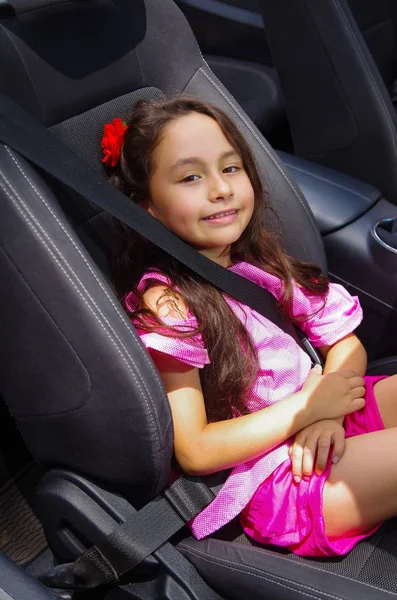 Close up of little beatiful girl sitting in the car wearing a pink dress and a red flower in her head using a safety belt