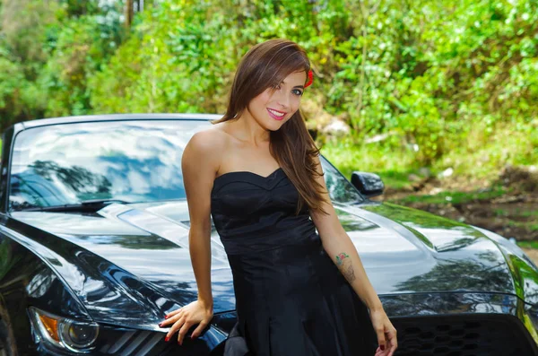 Close up of beautiful woman wearing a black dress and a red flower in her head and posing in front of a luxury black car on a road trip, the car standing on the sidelines, in a blurred nature — Stock Photo, Image