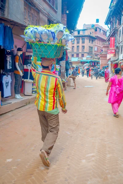 BHAKTAPUR, NEPAL - 04 DE NOVIEMBRE DE 2017: Un hombre no identificado cargando sobre su cabeza una canasta con cucharas al aire libre del antiguo edificio cerca de los mercados, con turistas caminando por Bhaktapur, Nepal —  Fotos de Stock