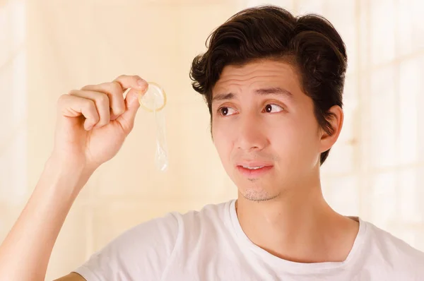Крупним планом тривожний чоловік тримає відкритий презерватив однією рукою перед обличчям, ознакою сексуальних захворювань, на розмитому фоні — стокове фото