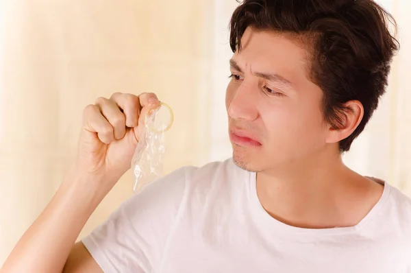 Porträtt av stilig man hålla en öppen kondom med ena handen och lukta framför hans ansikte, gör en äckligt ansikte, ett tecken på sex sjukdom, i en suddig bakgrund — Stockfoto