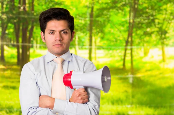 Portrait of a handsome mad man holding in his hand a megaphone with his arms crossed, in a blurred green background — Stock Photo, Image