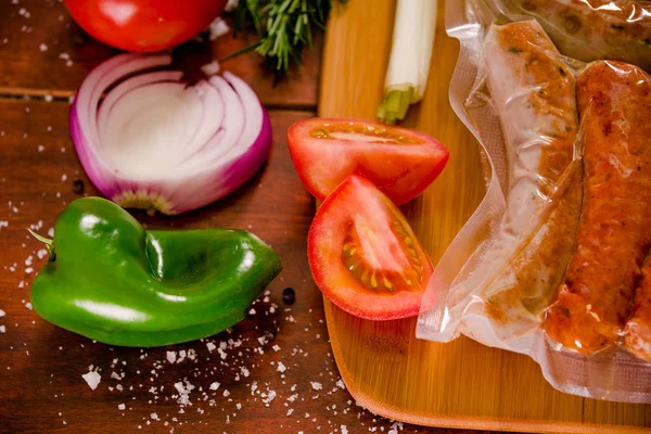 Above view of sausages inside of a plastic bags and vegetables, tomato, garlic, green pepper and onion and rosemary spices on a wooden background in rustic style — Stock Photo, Image