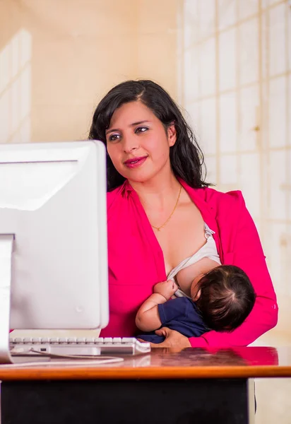 Jonge mooie zakenvrouw werken in het kantoor tijdens de vergadering met zijn babyjongen op haar werkplek, borstvoeding in een onscherpe achtergrond — Stockfoto