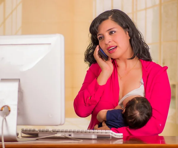 Junge schöne Geschäftsfrau, die im Büro arbeitet, während sie mit seinem kleinen Jungen an ihrem Arbeitsplatz sitzt und stillt, während sie ihr Handy vor verschwommenem Hintergrund benutzt — Stockfoto