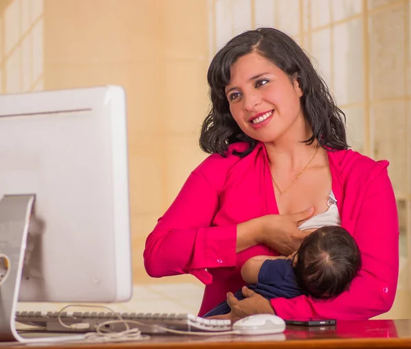 Jovem mulher de negócios bonita trabalhando no escritório enquanto se senta com seu menino em seu local de trabalho, amamentando em um fundo turvo — Fotografia de Stock