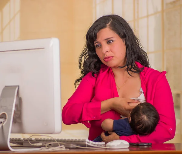 Junge schöne Geschäftsfrau, die im Büro arbeitet, während sie mit seinem kleinen Jungen an ihrem Arbeitsplatz sitzt und vor verschwommenem Hintergrund stillt — Stockfoto