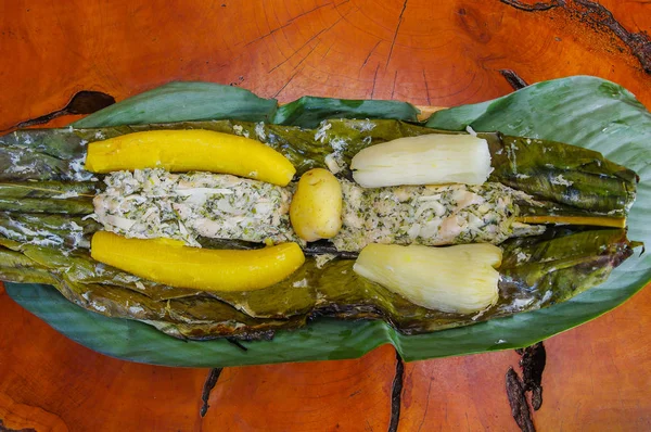 Sopra vista di deliziosi piatti tipici amazzonici, pesce cotto in foglia con yucca e piantaggine, servito in un piatto di legno su un tavolo di legno — Foto Stock