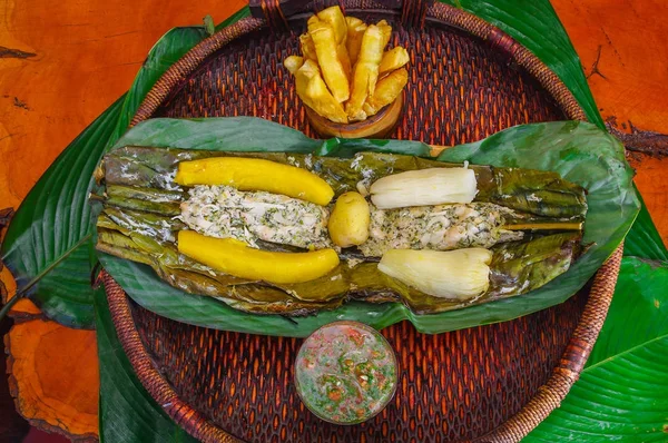 Above view of delicious typical amazonian food, fish cooked in a leaf with yucca and plantain, bowl of salad and fried yucca, served in a wooden plate over a wooden table — Stock Photo, Image