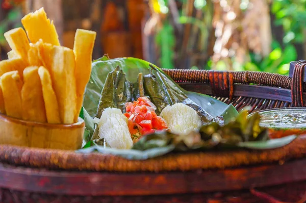Крупный план вкусной типичной амазонской пищи, рыба, приготовленная в листе с юккой и бананом, миска салата и жареной юкки, подается в деревянной тарелке над деревянным столом — стоковое фото