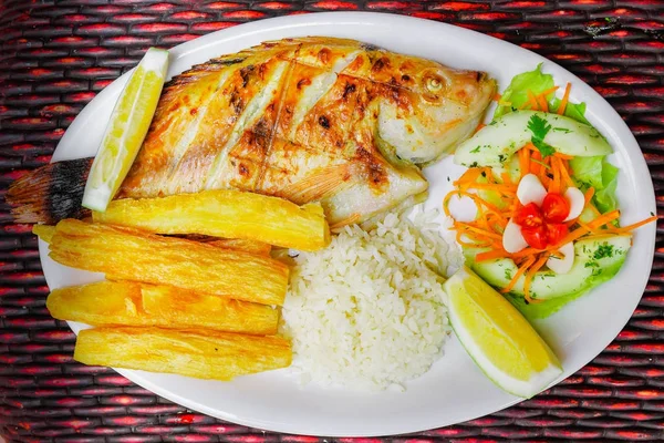 Vista superior del pescado frito servido con ensalada, arroz y yuca servidos en un plato blanco sobre una mesa de madera —  Fotos de Stock
