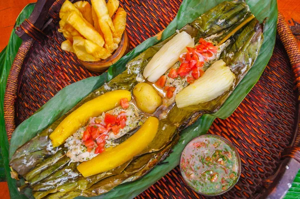 Vista superior de la deliciosa comida típica amazónica, pescado cocinado en una hoja con yuca y plátano, tazón de ensalada y yuca frita, servido en un plato de madera sobre una mesa de madera — Foto de Stock