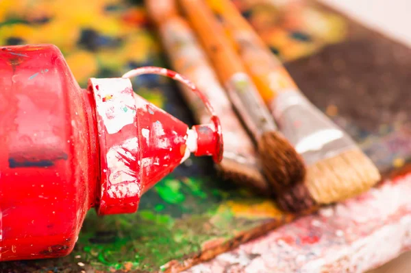 Close up de pincéis de tinta com uma cor de plástico vermelho sobre uma paleta de cores em um estúdio, em um fundo embaçado — Fotografia de Stock