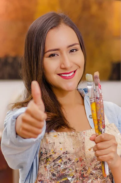 Güzel genç kadın ressam elinde boya fırçası ve içinde ona bir başparmak, bulanık bir arka planda işaret — Stok fotoğraf