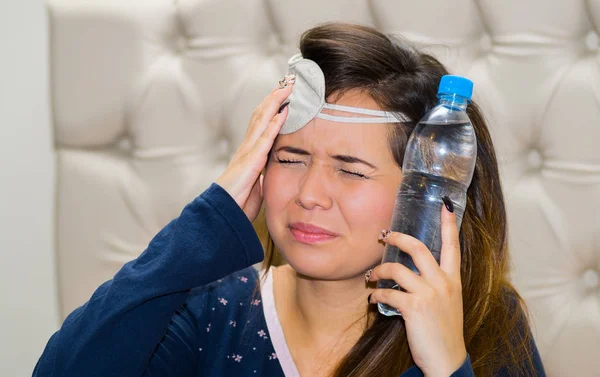 Donna ubriaca con una maschera per gli occhi addormentata in testa e con una bottiglia d'acqua che le preme in testa con l'altra mano, concetto di sbornia — Foto Stock