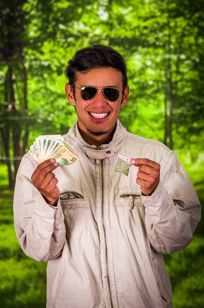 Detail prodejce muž nosí tmavé slunečních brýlí a drží v rukou peníze a drogy v jeho druhé, obchodování s drogami, zločinem, rozmazané pozadí — Stock fotografie
