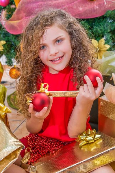 Close up van lachende meisje draagt een rode blouse en holding een kerst bal, decoratie en cadeau in haar handen, met een kerstboom achter, Kerstmis concept — Stockfoto