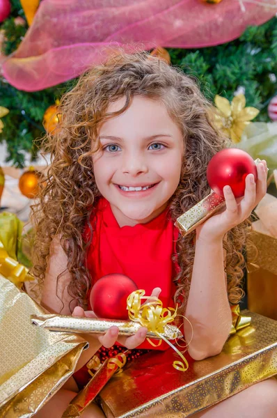 Close up van lachende meisje draagt een rode blouse en holding een kerst bal en cadeau in elke handen, met een kerstboom achter, Kerstmis concept — Stockfoto