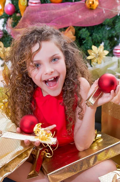 Close up de menina sorridente vestindo uma blusa vermelha e segurando uma bola de Natal e presente em cada mãos, com uma árvore de Natal atrás, conceito de natal — Fotografia de Stock