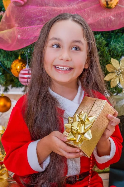 Close up van lachende meisje draagt een rode santa kostuum en bedrijf van een gift in haar handen, met een kerstboom achter, Kerstmis concept — Stockfoto