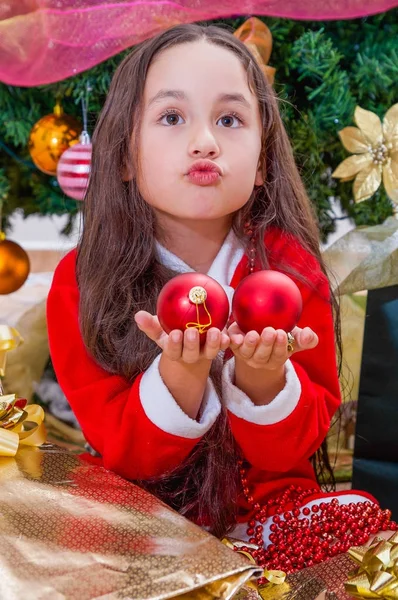 Close up de menina feliz vestindo um traje de Papai Noel vermelho e segurando duas bolas de Natal em suas mãos fazendo cara engraçada, com uma árvore de Natal atrás, conceito de natal — Fotografia de Stock