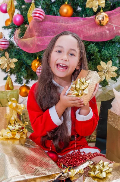 Primo piano di sorridente ragazza con un costume da Babbo Natale rosso e tenendo un regalo in mano, con un albero di Natale dietro, concetto di Natale — Foto Stock