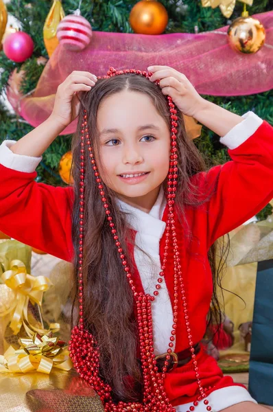 Close up van gelukkig meisje draagt een rode jurk en houdt een Kerstdecoratie in haar handen over haar hoofd, met een kerstboom achter, Kerstmis concept — Stockfoto