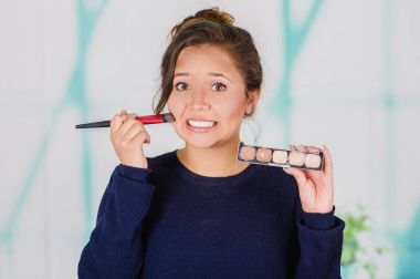 Close up of young beautiful woman holding a make up palette and doing crazy make-up in her face using a brush, in a blurred background clipart