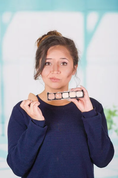 Acercamiento de una joven preocupada sosteniendo una paleta de maquillaje y haciendo maquillaje loco en su cara usando una esponja, en un fondo borroso —  Fotos de Stock