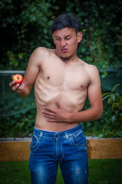 Primer plano del hombre solitario que sufre de anorexia, sufriendo dolor en el estómago cuando mira una manzana y haciendo caras repugnantes, en un fondo borroso — Foto de Stock