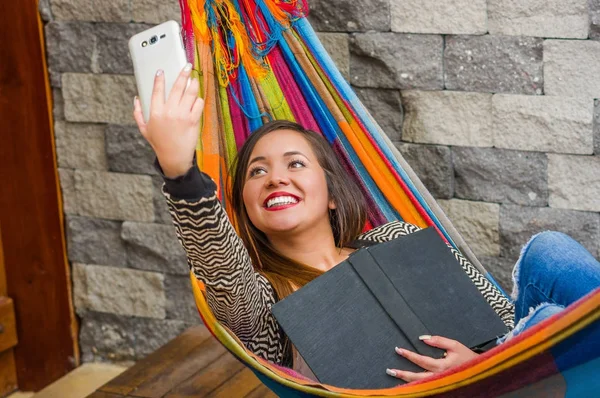 Primer plano de la joven hermosa mujer relajándose en una hamaca y tomando una selfie con su tableta mientras sostiene un libro con su otra mano, fondo borroso — Foto de Stock