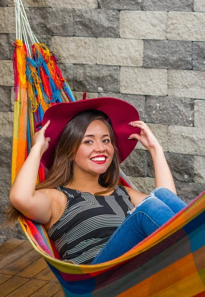 Acercamiento de la mujer hermosa joven que relaja en una hamaca y que usa un sombrero rojo de la manera que sostiene con ambas manos, en fondo borroso — Foto de Stock