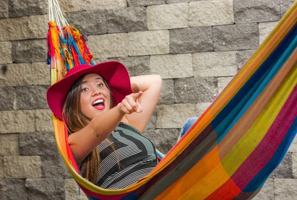 Gros plan de jeune belle femme portant un chapeau de mode rouge et se relaxant dans un hamac, pointant quelque part avec son bras, dans un fond flou — Photo